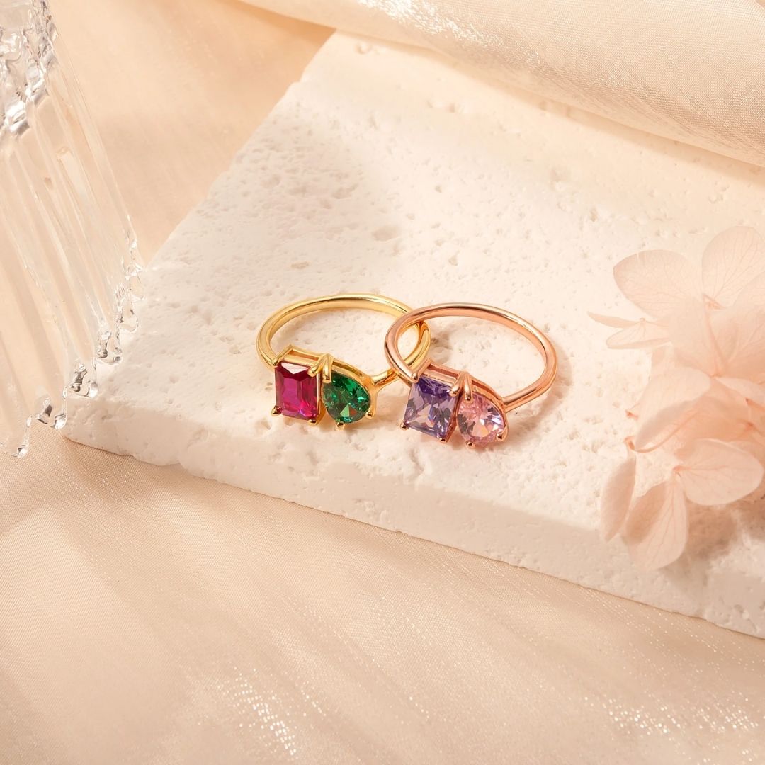 Dos anillos de oro "Anillo Eterno con piedras de nacimiento" (Oferta 2x1) con coloridas piedras de nacimiento se exhiben sobre una superficie de textura blanca con tela transparente y flores pálidas, constituyendo un encantador regalo para parejas.