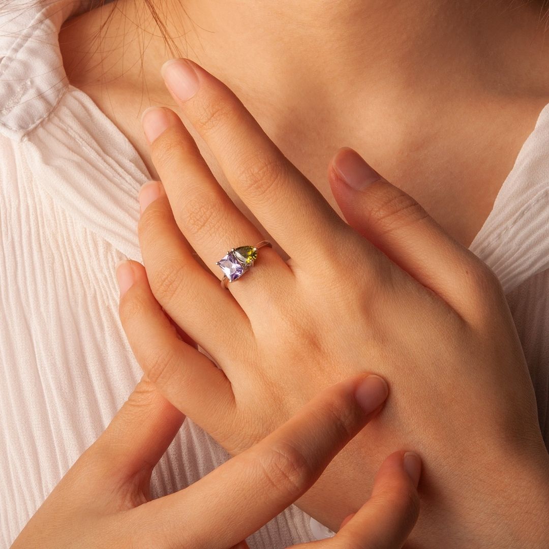 Una persona con una blusa blanca plisada toca suavemente su mano, mostrando el Anillo Eterno con piedras de nacimiento (Oferta 2x1) con una gema grande de color morado claro y un pequeño detalle verde, ideal como regalo para parejas.