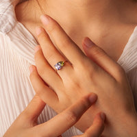 Una persona con una blusa blanca plisada toca suavemente su mano, mostrando el Anillo Eterno con piedras de nacimiento (Oferta 2x1) con una gema grande de color morado claro y un pequeño detalle verde, ideal como regalo para parejas.