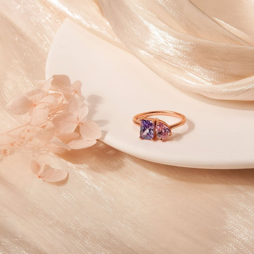 El Anillo Eterno con piedras de nacimiento (Oferta 2x1), en oro, con dos gemas-una morada y una rosa-simbolizando meses de nacimiento, descansa sobre un plato blanco entre flores claras y tela beige brillante.