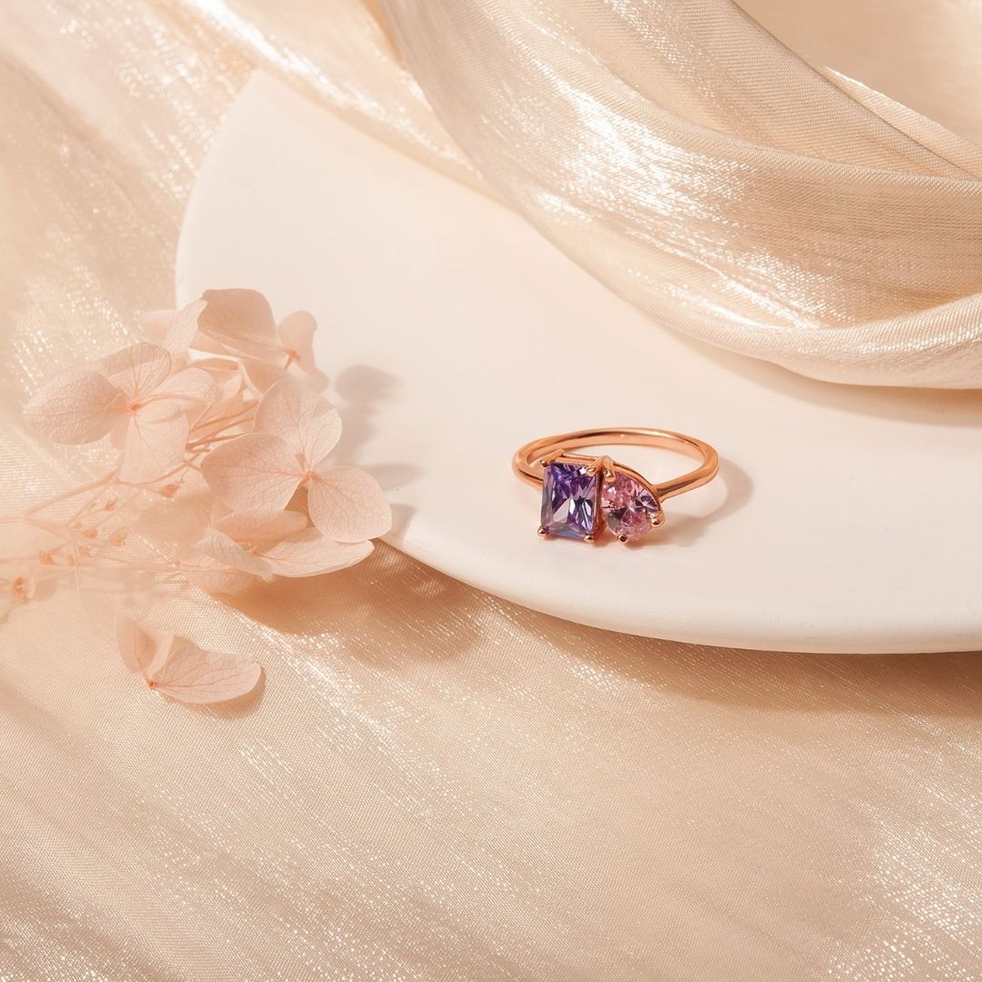 El Anillo Eterno con piedras de nacimiento (Oferta 2x1), en oro, con dos gemas-una morada y una rosa-simbolizando meses de nacimiento, descansa sobre un plato blanco entre flores claras y tela beige brillante.