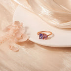 El Anillo Eterno con piedras de nacimiento (Oferta 2x1), en oro, con dos gemas-una morada y una rosa-simbolizando meses de nacimiento, descansa sobre un plato blanco entre flores claras y tela beige brillante.