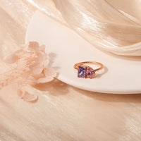 El Anillo Eterno con piedras de nacimiento (Oferta 2x1), en oro, con dos gemas-una morada y una rosa-simbolizando meses de nacimiento, descansa sobre un plato blanco entre flores claras y tela beige brillante.