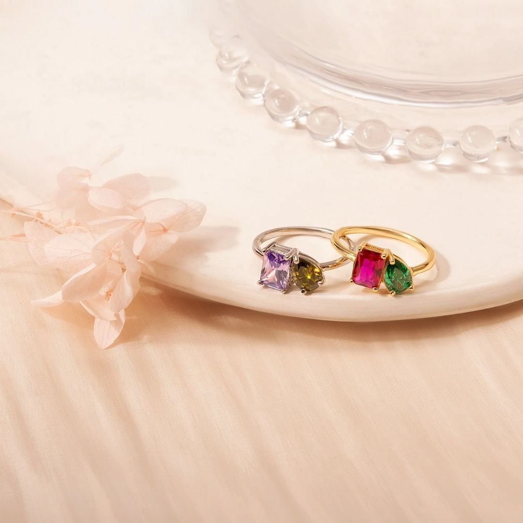 Dos Anillos Eternos con piedras de nacimiento (Oferta 2x1), uno plateado con gemas morada y verde olivo, otro dorado con piedras rosa y verde, se exhiben sobre un plato crema junto a flores secas: el regalo ideal para parejas.
