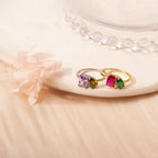 Dos Anillos Eternos con piedras de nacimiento (Oferta 2x1), uno plateado con gemas morada y verde olivo, otro dorado con piedras rosa y verde, se exhiben sobre un plato crema junto a flores secas: el regalo ideal para parejas.