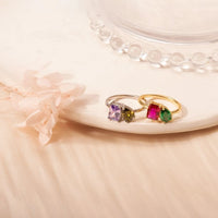 Dos Anillos Eternos con piedras de nacimiento (Oferta 2x1), uno plateado con gemas morada y verde olivo, otro dorado con piedras rosa y verde, se exhiben sobre un plato crema junto a flores secas: el regalo ideal para parejas.