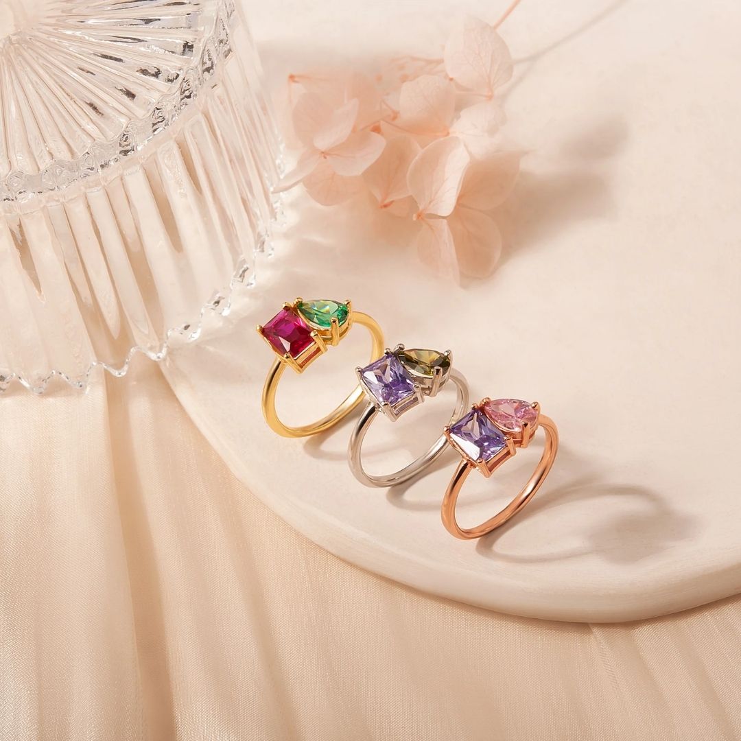 Cuatro Anillos Eternos con piedras de nacimiento (Oferta 2x1) se exhiben sobre una suave superficie, junto a un recipiente de vidrio y flores secas, creando un ambiente delicado y sofisticado.