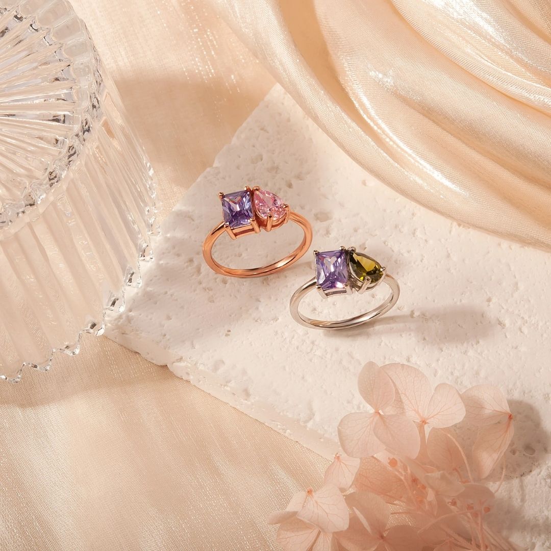 Dos Anillos Eternos con piedras de nacimiento (Oferta 2x1): uno con gemas moradas y rosas en oro rosa, otro con piedras moradas y verdes en plata. Un regalo ideal para parejas, presentado entre telas suaves y flores.