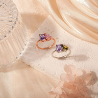 Dos Anillos Eternos con piedras de nacimiento (Oferta 2x1): uno con gemas moradas y rosas en oro rosa, otro con piedras moradas y verdes en plata. Un regalo ideal para parejas, presentado entre telas suaves y flores.