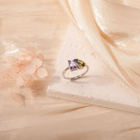 Un Anillo Eterno de plata con piedras de nacimiento (Oferta 2x1) con una piedra morada y otra verde, mostradas sobre una superficie blanca texturizada con tela transparente y flores secas de color rosa pálido-un regalo ideal para parejas.