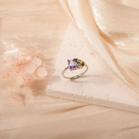 Un Anillo Eterno de plata con piedras de nacimiento (Oferta 2x1) con una piedra morada y otra verde, mostradas sobre una superficie blanca texturizada con tela transparente y flores secas de color rosa pálido-un regalo ideal para parejas.