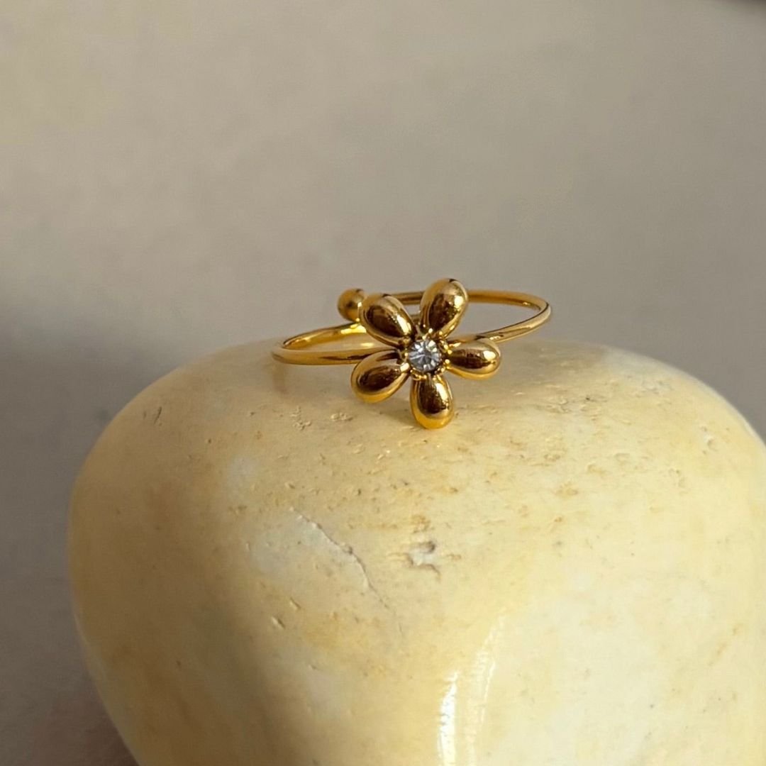 A joya delicada, el Anillo Flor ajustable en oro destaca con un pequeño diamante central y reposa elegantemente sobre una piedra clara, todo en un fondo neutro.