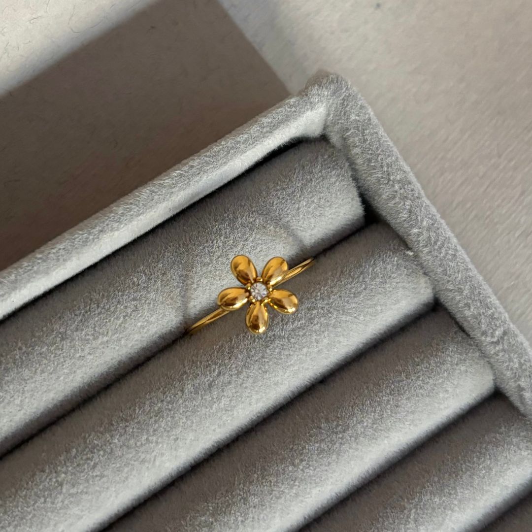 El Anillo Flor ajustable es un anillo de oro con una flor en forma de margarita con una piedra preciosa central, presentado en una caja de terciopelo gris claro-una verdadera joya delicada.