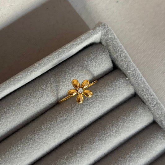 El Anillo Flor ajustable es un anillo de oro con una flor en forma de margarita con una piedra preciosa central, presentado en una caja de terciopelo gris claro-una verdadera joya delicada.
