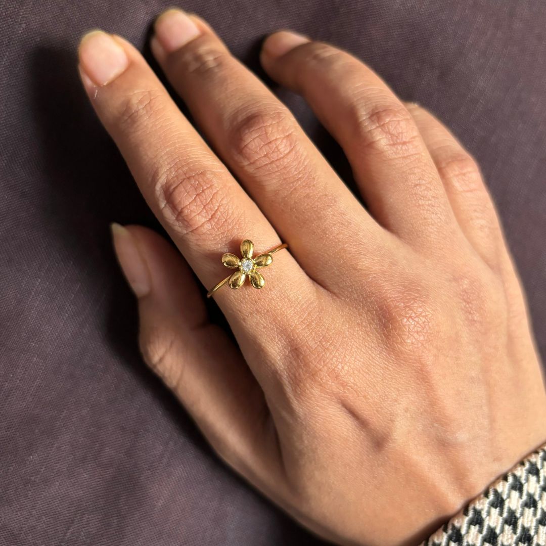 Una mano con uñas bien cuidadas luce el Anillo Flor ajustable, una joya delicada con oro y una gema verde en el centro, reposando sobre una superficie púrpura oscura.