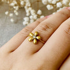 Una mano luce el Anillo Flor ajustable, una joya delicada con diseño de flor y diamante central, sobre un fondo suave adornado con flores blancas secas.