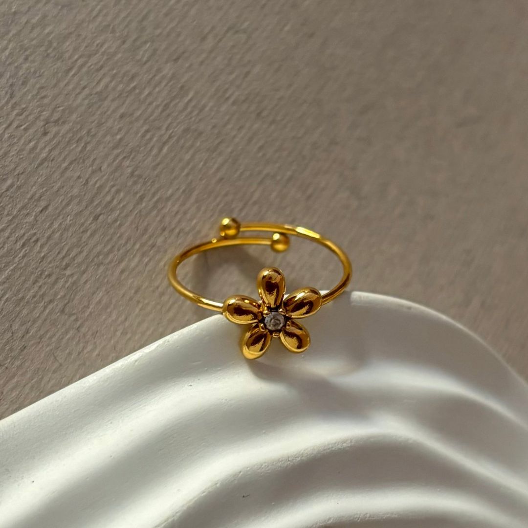 La delicada joya Anillo Flor ajustable, en dorado y con pequeña gema central, descansa sobre una superficie blanca texturizada.
