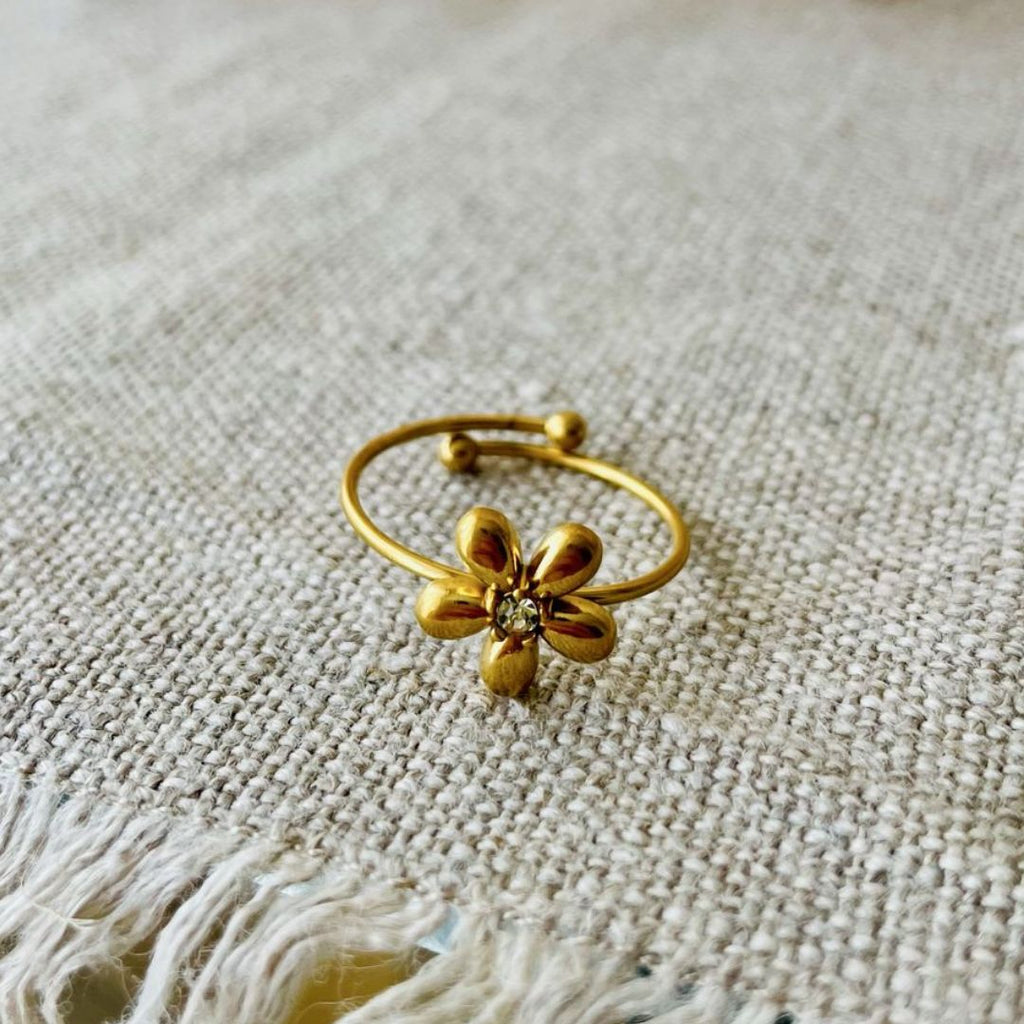 El Anillo Flor ajustable, un delicado anillo dorado en forma de flor con una gema central, descansa sobre un tejido beige texturizado-una joya perfecta para cualquier ocasión.