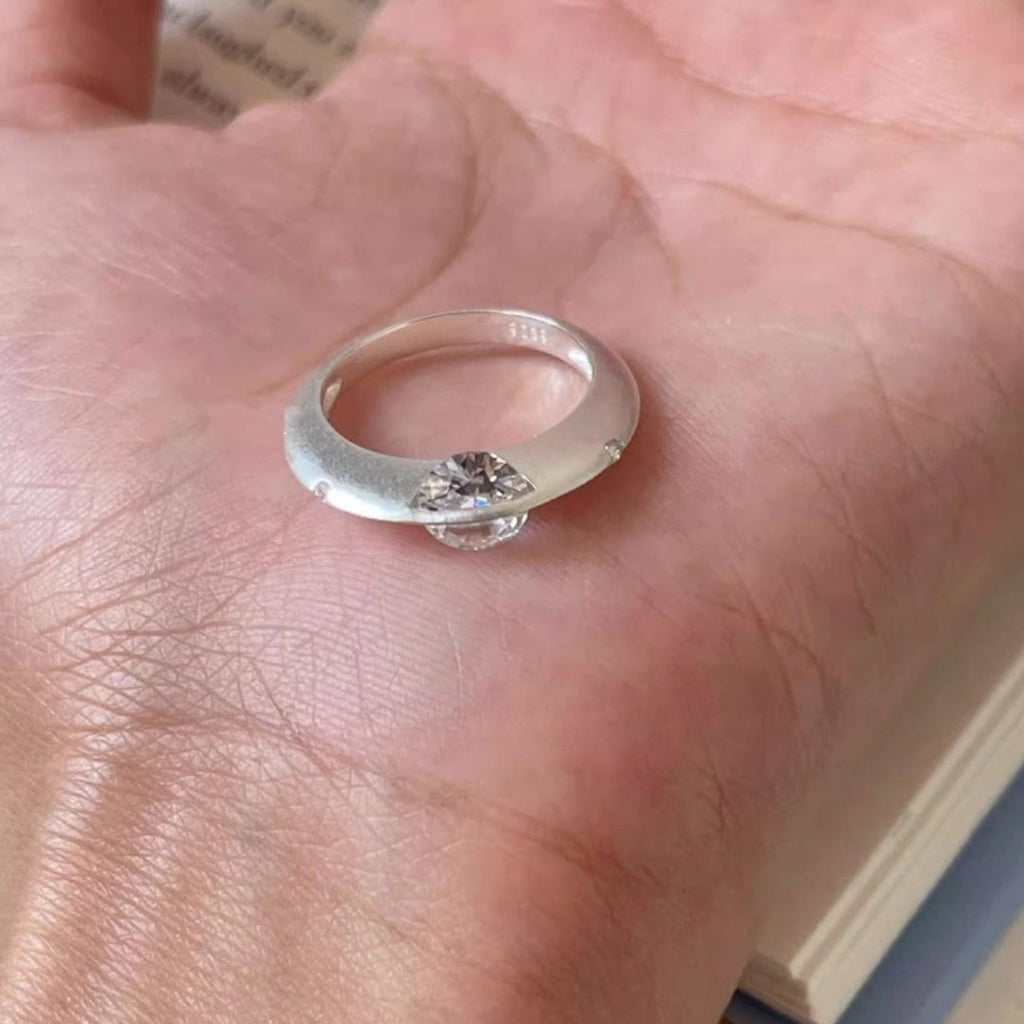 Primer plano de una mano que sostiene un Anillo giratorio estelar con una gran piedra preciosa transparente en el centro, sobre la palma. El fondo muestra el borde de un libro abierto.
