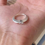 Primer plano de una mano que sostiene un Anillo giratorio estelar con una gran piedra preciosa transparente en el centro, sobre la palma. El fondo muestra el borde de un libro abierto.