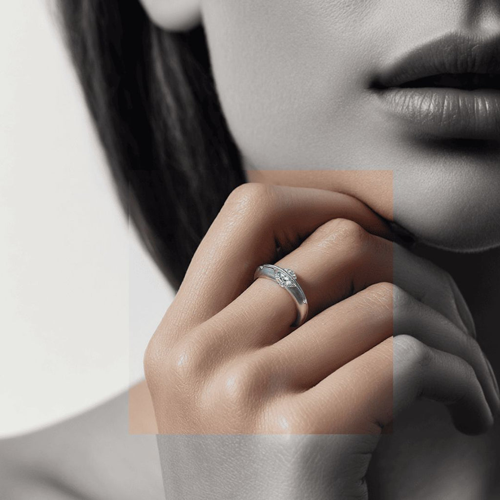 Un primer plano muestra la mano de una mujer cerca de la barbilla, destacando el Anillo giratorio estelar, un anillo ajustable de acero quirúrgico blanco con una pequeña piedra preciosa en el dedo anular. La mayoría de la foto es en blanco y negro, salvo la mano en color.