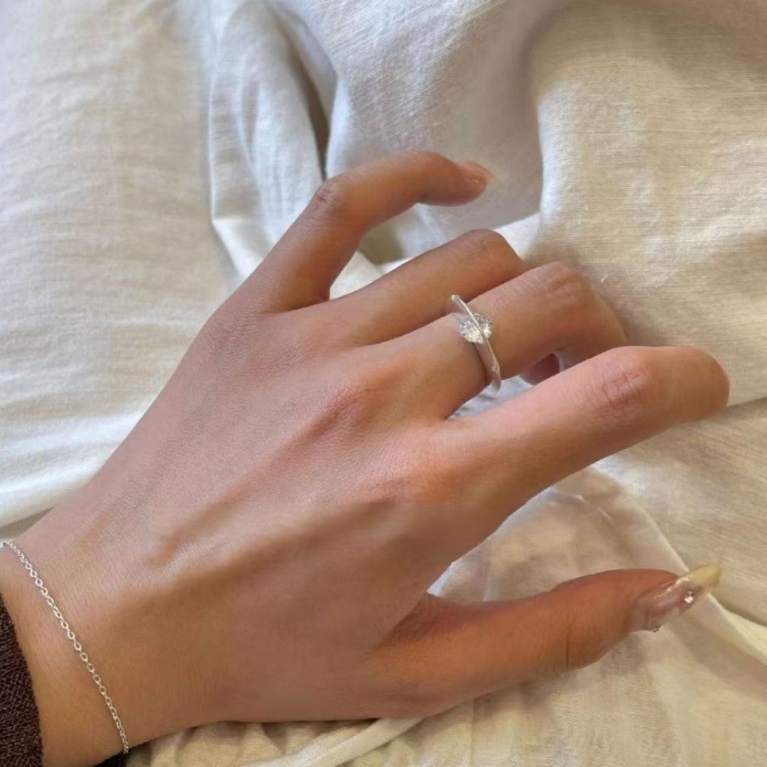 Una mano adornada con el Anillo giratorio estelar y una delicada pulsera de plata descansa sobre una tela suave de tonos claros. Las uñas están cuidadas, con una uña del meñique notablemente más larga y decorada que añade un toque elegante.