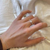 Una mano adornada con el Anillo giratorio estelar y una delicada pulsera de plata descansa sobre una tela suave de tonos claros. Las uñas están cuidadas, con una uña del meñique notablemente más larga y decorada que añade un toque elegante.
