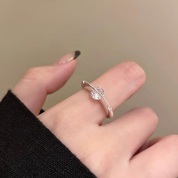 Primer plano de una mano que lleva el Anillo giratorio estelar, un anillo ajustable de acero quirúrgico blanco con dos gemas redondas transparentes separadas por una barra curva. Las uñas de la persona están desnudas con las puntas negras.