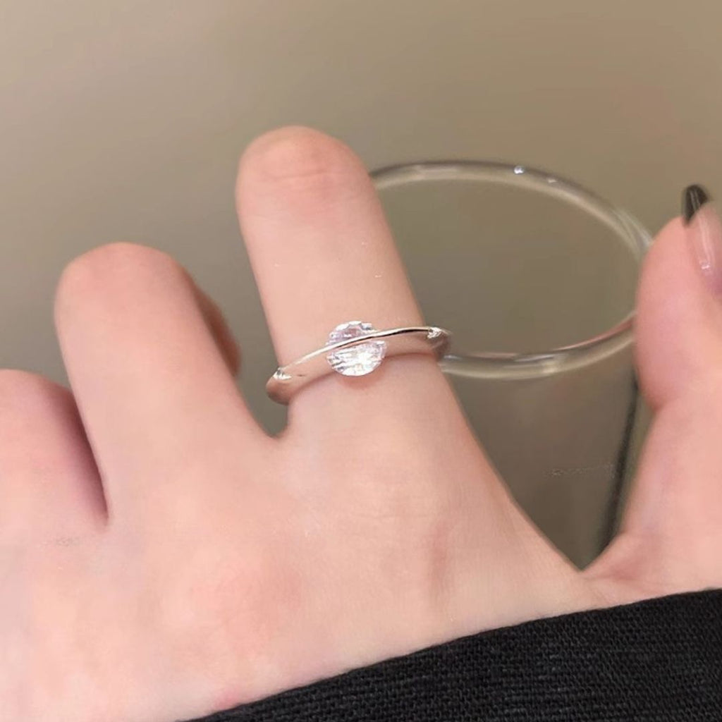 Una mano lleva el Anillo giratorio estelar, un anillo minimalista ajustable de acero quirúrgico blanco con una gema redonda transparente. La persona sostiene un vaso transparente sobre un fondo neutro.