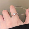 Una mano lleva el Anillo giratorio estelar, un anillo minimalista ajustable de acero quirúrgico blanco con una gema redonda transparente. La persona sostiene un vaso transparente sobre un fondo neutro.