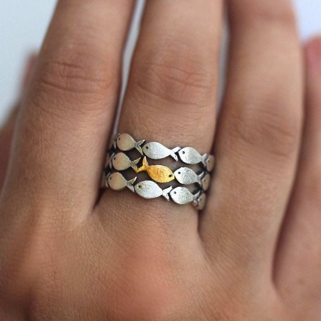 Primer plano de una mano que lleva el Anillo "Nada contra la corriente", un anillo de acero inoxidable con motivos de peces plateados y un destacado pez dorado en el centro.