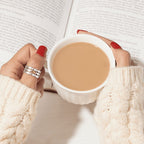 Una persona con las uñas pintadas de rojo y el anillo "Nada contra la corriente" sostiene una taza de café o té, con un jersey de punto color crema, con un libro abierto al fondo.