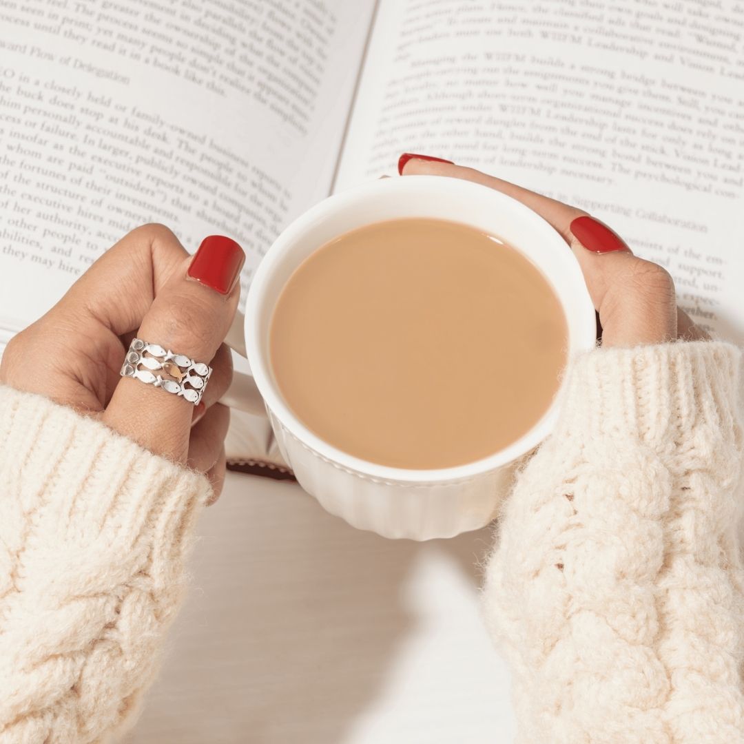 Una persona con las uñas pintadas de rojo y el anillo "Nada contra la corriente" sostiene una taza de café o té, con un jersey de punto color crema, con un libro abierto al fondo.