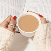 Cargar imagen en el visor de la galería, Una persona con las uñas pintadas de rojo y el anillo &quot;Nada contra la corriente&quot; sostiene una taza de café o té, con un jersey de punto color crema, con un libro abierto al fondo.