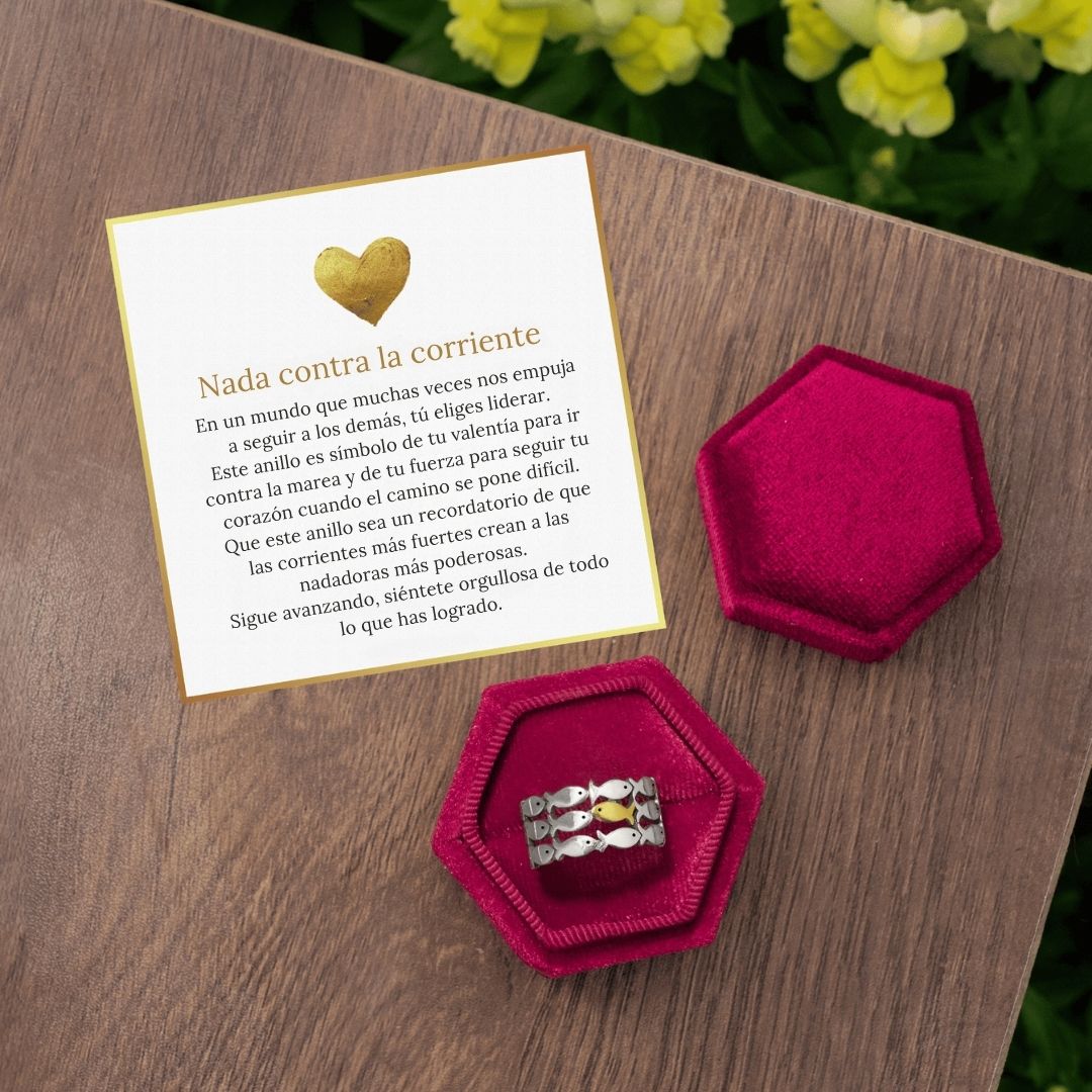 El Anillo "Nada contra la corriente", de plata y diseño de ola, reposa en una caja hexagonal rosa sobre madera. A su lado hay una tarjeta en español con un corazón dorado y mensaje inspirador, con flores amarillas al fondo.