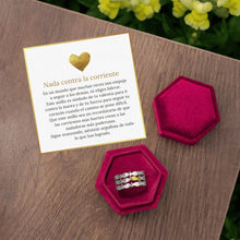 Cargar imagen en el visor de la galería, El Anillo &quot;Nada contra la corriente&quot;, de plata y diseño de ola, reposa en una caja hexagonal rosa sobre madera. A su lado hay una tarjeta en español con un corazón dorado y mensaje inspirador, con flores amarillas al fondo.