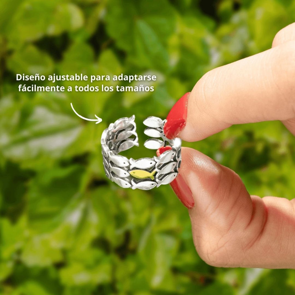 Una mano con esmalte rojo sostiene el anillo "Nada contra la corriente", un anillo ajustable de acero inoxidable en forma de hojas enlazadas, sobre fondo verde. Texto: "Diseño ajustable para adaptarse fácilmente a todos los tamaños.
