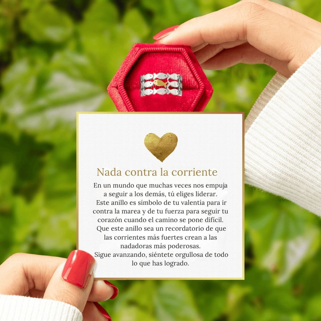 Una persona con las uñas pintadas de rojo sostiene el Anillo "Nada contra la corriente" en una caja roja sobre una tarjeta con un mensaje motivador en español y un corazón dorado, sobre hojas verdes.
