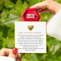 Una persona con las uñas pintadas de rojo sostiene el Anillo "Nada contra la corriente" en una caja roja sobre una tarjeta con un mensaje motivador en español y un corazón dorado, sobre hojas verdes.