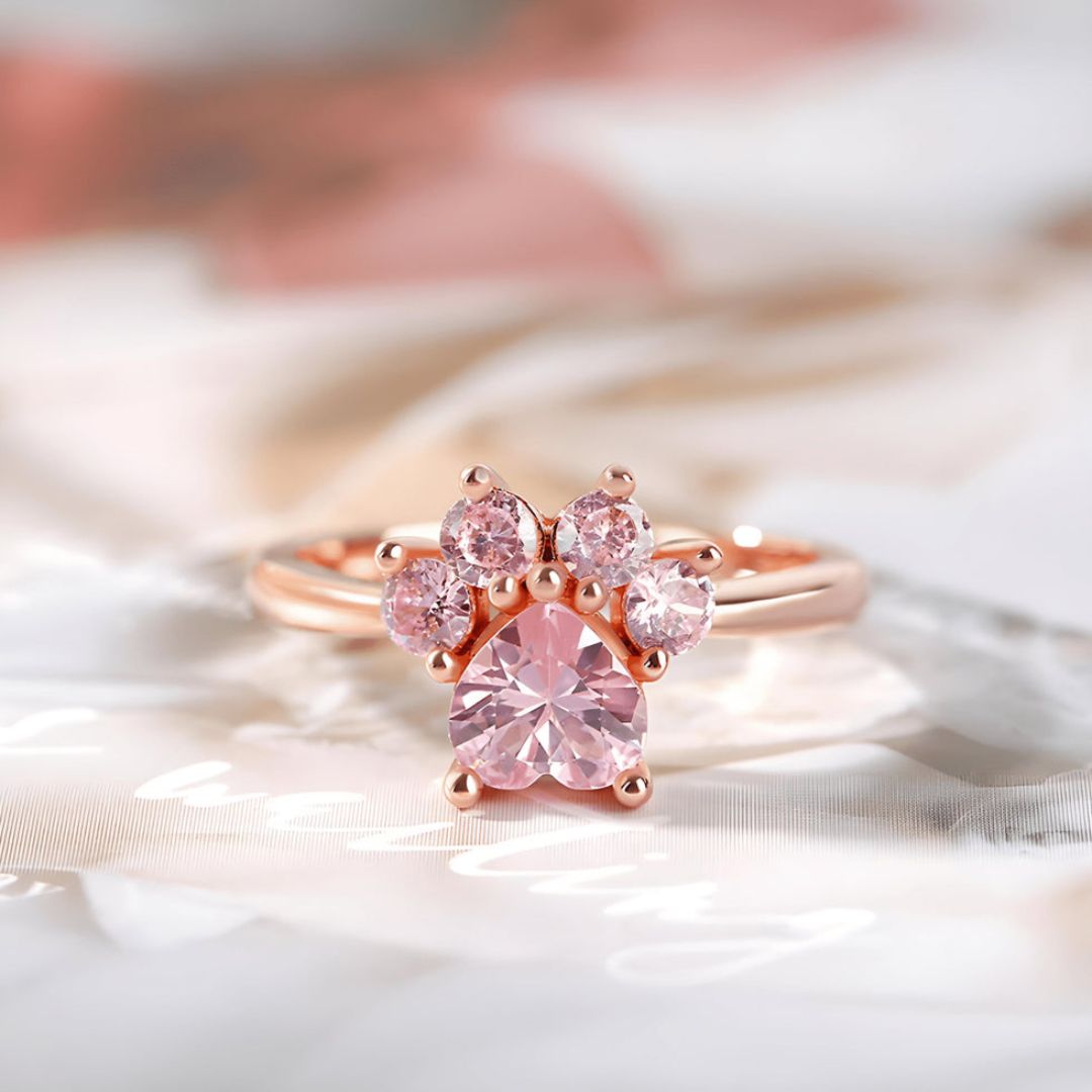 El Anillo Patita rosa en dorado muestra cinco piedras preciosas rosas dispuestas como una huella de pata, lo que lo convierte en un regalo ideal para amantes de mascotas, sobre un fondo suave de color pastel.