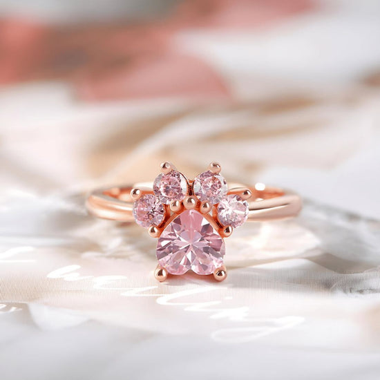 El Anillo Patita rosa en dorado muestra cinco piedras preciosas rosas dispuestas como una huella de pata, lo que lo convierte en un regalo ideal para amantes de mascotas, sobre un fondo suave de color pastel.