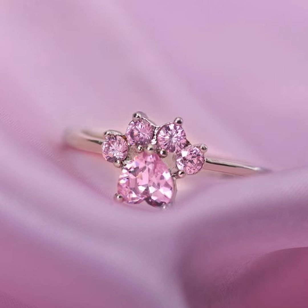 El Anillo Patita rosa es un delicado anillo de plata adornado con cinco gemas rosas en forma de pata, que se muestra sobre una suave tela rosa: perfecto como regalo para los amantes de las mascotas.