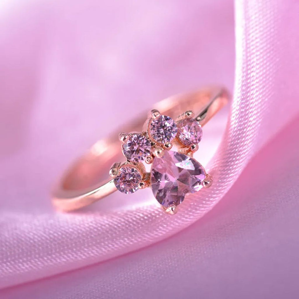 El Anillo Patita rosa, con gema central en forma de corazón y cuatro piedras rosas, descansa sobre suave satén rosado; es el recuerdo de mascota perfecto o un regalo ideal para amantes de mascotas.