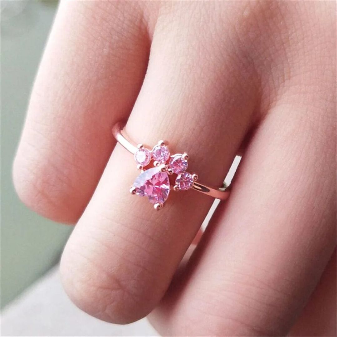 Una mano luce el Anillo Patita rosa, un delicado anillo de oro adornado con gemas rosas en forma de flor o mariposa, perfecto como regalo para los amantes de las mascotas.