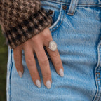 Una mano con uñas pálidas y cuidadas descansa sobre el bolsillo de unos vaqueros azul claro, y lleva un gran Anillo plateado personalizado con foto con la letra M. La persona también lleva un jersey marrón, a cuadros y peludo.