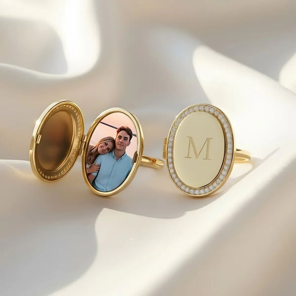 Anillo personalizado con foto: un anillo ovalado dorado con borde de perlas y la letra M grabada, que se abre para mostrar una foto de una pareja sonriente. Ideal como regalo personalizado y presentado sobre tela clara y suave.