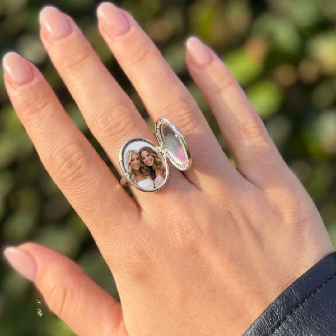 Una mano lleva un Anillo personalizado con foto que se abre para mostrar una pequeña foto de dos mujeres sonrientes en su interior. El fondo verde difuminado hace de este Anillo personalizado con foto un regalo personalizado ideal.