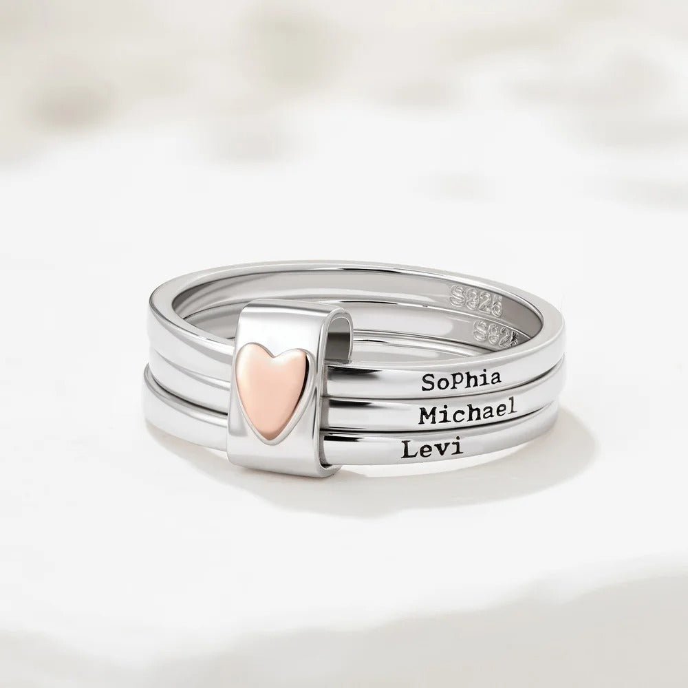 El Anillo personalizado con nombres presenta tres aros de acero inoxidable apilados con un dije de corazón en oro rosa y lleva grabados los nombres Sophia, Michael y Levi. Se muestran sobre una superficie blanca y texturizada.