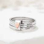 El Anillo personalizado con nombres presenta tres aros de acero inoxidable apilados con un dije de corazón en oro rosa y lleva grabados los nombres Sophia, Michael y Levi. Se muestran sobre una superficie blanca y texturizada.