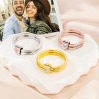 Three Anillos personalizados con nombres-uno en plata, uno en oro y uno en oro rosa de acero inoxidable con un pequeño corazón-se exhiben sobre un plato en forma de corazón; al fondo se ve la foto sonriente de una pareja.